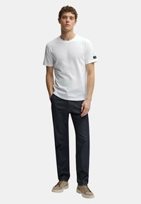 T-shirt bianca a maniche corte con collo rotondo, abbinata a jeans blu scuro e scarpe beige con lacci, con un'etichetta logo sulla manica.