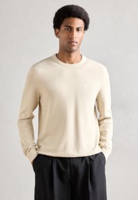 Jersey beige de punto con cuello redondo y mangas largas, con un acabado texturizado sutil. Combinado con pantalones oscuros.