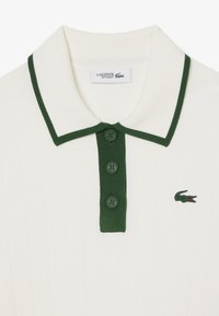 Witte geribbelde poloshirt met een groene kraag en drie bijpassende knopen. Heeft een klein groen geborduurd krokodillenlogo aan de linkerkant.