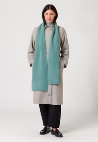 Cappotto di lana grigio con design a cintura, abbinato a una sciarpa in teal a coste. Pantaloni neri a gamba larga e mocassini neri completano l'outfit.