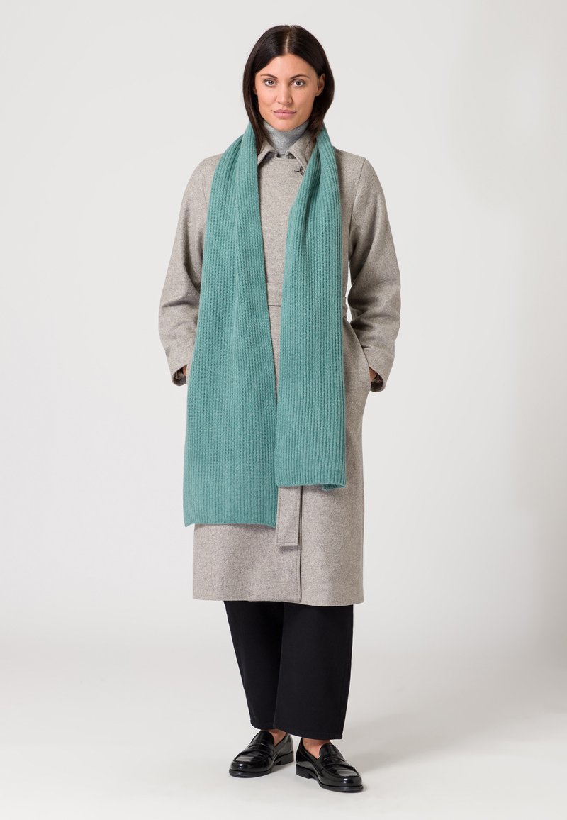 Cappotto di lana grigio con design a cintura, abbinato a una sciarpa in teal a coste. Pantaloni neri a gamba larga e mocassini neri completano l'outfit.