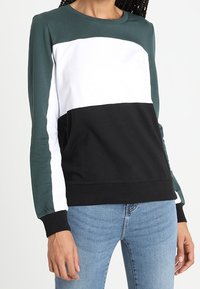 Färgblockad sweatshirt med mörkgrön, vit och svart sektioner. Tillverkad av ett mjukt tyg med långa ärmar och ribbade cuffs.