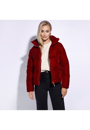 Rode fluwelen pufferjack met een hoge kraag, ritssluiting en elastische manchetten. Draagt over een lichtgekleurde top en zwarte broek. Glad textuur.