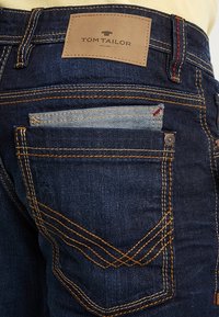 Jeans de mezclilla azul oscuro con una etiqueta de cuero marrón etiquetada "TOM TAILOR", con costuras de contraste y un bolsillo con un inserto de mezclilla más clara.
