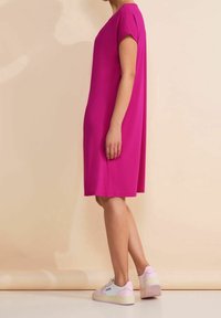 Kvinna som står på sidan iförd en knälång magentafärgad klänning med korta ärmar och vita och rosa sneakers mot en beige bakgrund.