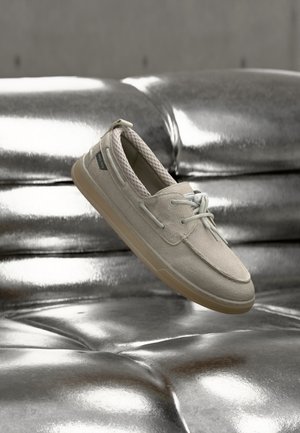 Beige ruskind sneakers med snørebånd, der svæver over en skinnende sølv tuftet lædersofa.