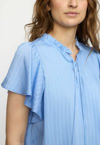 Blusa azzurro chiaro realizzata in tessuto texture con un motivo a righe verticali. Presenta un colletto arricciato e maniche corte a volant; chiusura con bottone.