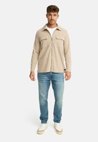 Mand iført en beige lynlåsjakke, hvid t-shirt, blå jeans og hvide sneakers, der står foran en ensfarvet baggrund.