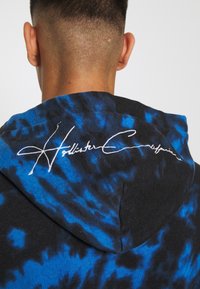 Černá mikina s kapucí s modrým tie-dye vzorem, na zadní straně kapuce je bílý vyšívaný logo "Hollister California".