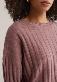 Strak aansluitende, geribbelde trui van zacht mauve stof; ronde halslijn en iets verlaagde schouders. De textuur is glad met een lichte uitstraling.