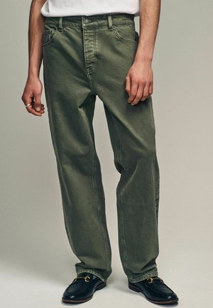 Grüne Baumwollhose mit lockerer Passform, ausgestattet mit fünf Taschen, Gürtelschlaufen und einem Knopfverschluss, kombiniert mit schwarzen Slippern.