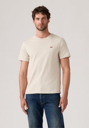 Man in een beige Levi's t-shirt en blauwe spijkerbroek die tegen een lichte, effen achtergrond staat.