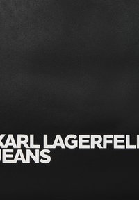 Černý texturovaný povrch s tučnými bílými velkými písmeny, která čtou "KARL LAGERFELD JEANS" a jsou částečně viditelná ve spodní části.
