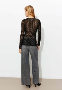 Haut en dentelle noire à manches longues, avec un motif floral et un dos transparent, associé à un jean gris taille haute à jambes larges et des talons noirs.