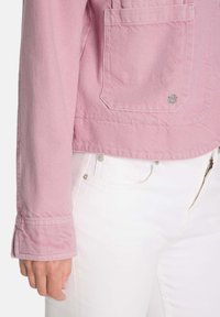 Rosa kurze Jacke aus strukturiertem Stoff, mit einer großen Fronttasche, silbernen Hardware-Details und breiten Manschetten. Kombiniert mit weißen Hosen.