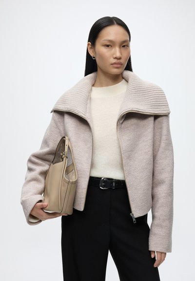 Femme aux cheveux noirs et raides portant une veste zippée beige clair, un pull crème, un pantalon noir, tenant un sac à main beige sur fond uni.
