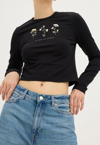 Top negro de manga larga y cropped con gráficos florales en blanco y amarillo, con el texto "sin lluvia: no hay flores" debajo, combinado con unos jeans ajustados de cintura alta en azul.