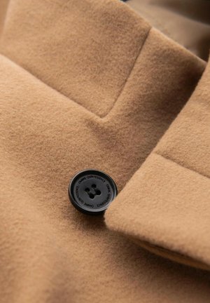 Kamelí vlna s texturovaným povrchem, která má na klopě černý čtyřdírkový knoflík a elegantní design límce.
