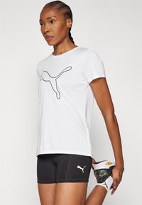 Valge spordit-särk musta Puma logoga, mille juurde on sobitatud mustad lühikesed püksid valge logoga. Mudel kannab musti ja valgeid saapaid.