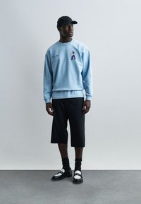 Άνδρας με ανοιχτό μπλε φούτερ με σχέδιο, μαύρο καπέλο, μαύρα σορτς μέχρι το γόνατο, μαύρες κάλτσες και ασπρόμαυρα μοκασίνια, στέκεται δίπλα σε λευκό τοίχο.