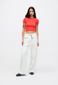 Κόκκινο cropped t-shirt με μαύρο λογότυπο, συνδυασμένο με ανοιχτογκρι φόρμα με ρυθμιζόμενο κορδόνι και πλαϊνή τσέπη, φορεμένο με μαύρα παπούτσια.