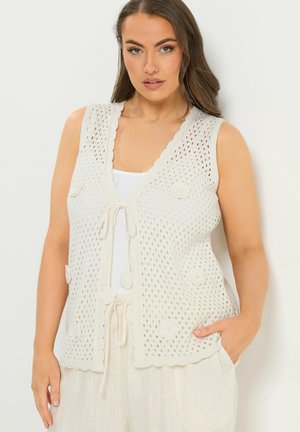 Mujer vistiendo un chaleco blanco de crochet sin mangas con detalles florales, atado al frente, sobre una blusa blanca y pantalones sueltos de color claro.