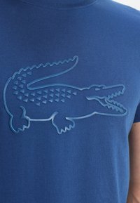 Blå bomullströja med en texturerad, reliefpräglad krokodilgrafik i en ljusare blå nyans, med en slät yta och rund halsringning.