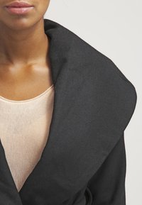 Cappotto in misto lana nero con un grande colletto strutturato, indossato sopra una maglia beige chiaro. La texture appare morbida e leggermente pelosa.