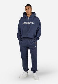 Mørkeblå hætte sweatshirt med lynlås, "PEGLADOR" logo i hvid, parret med matchende sweatpants og hvide sneakers. Glat bomuldsblandingsstof.
