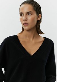 Melnas, pārmērīgs džemperis ar dziļu V veida kakla izgriezumu un ribota tekstūra. Audums izskatās mīksts un ikdienišķs, ar nokristām plecu šuvēm un brīvu piegriezumu.