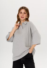 Grijze oversized polo met een kraag, voorzien van korte mouwen en een ontspannen pasvorm. Het materiaal lijkt zacht met een subtiele textuur.