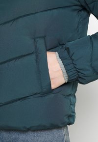 Polstret turkisjakke med et vandret quiltet design, med strikkede grå manchetter og en sidelomme. Blød, glat stoftekstur.