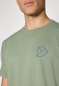 Žalios spalvos medvilnės marškinėliai su apvaliu iškirpimu. Kairėje krūtinės pusėje yra geometrinis mėlynas logotipo dizainas. Trumpomis rankovėmis ir minkšta tekstūra.