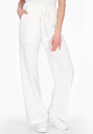 Person iført løse hvide bukser med snøre og hvide sneakers, med den ene hånd i lommen og den anden hvilende på låret.