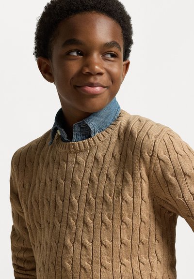 Beidzis kabels vītne džemperis ar klasisku apaļu kakla izgriezumu, uzvilkts pāri tumši zilai džinsu krekliņam. Faktūra ir bieza, ar blīvām, paaugstinātām rakstām.