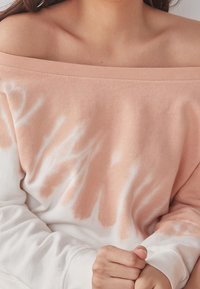 Přes oversized mikina s designem bez ramene, která má broskvové a bílé tie-dye vzory a jemnou bavlněnou texturu.