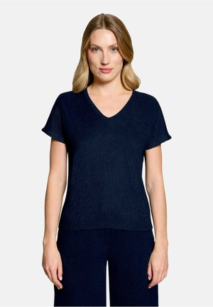 Kvinde med blondt hår iført en marineblå T-shirt med korte ærmer og V-hals samt matchende bukser, stående foran en ensfarvet lys baggrund.