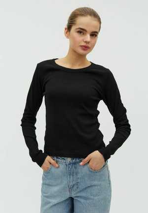 Mladá žena s vlasmi zopnutými dozadu, oblečená v čiernom tričku s dlhým rukávom a svetlomodrých džínsoch, ruky vreckách, neutrálne pozadie.