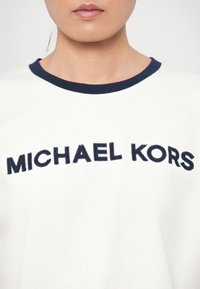 Maglietta bianca con colletto rotondo blu navy, caratterizzata da un audace testo ricamato "MICHAEL KORS" sul petto. Tessuto morbido.