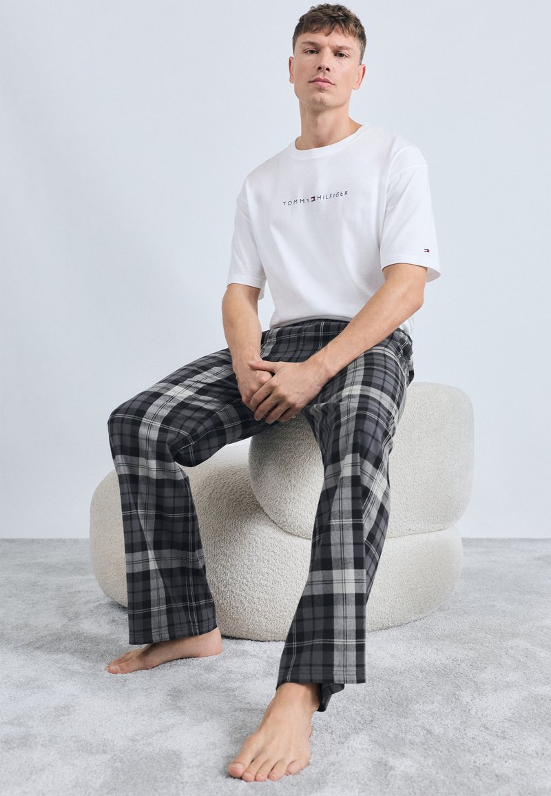 Fehér, rövid ujjú póló "TOMMY HILFIGER" logóval; szürke kockás otthoni nadrág fekete-fehér kockás mintával; mezítláb.