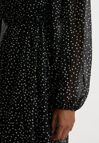 Schwarz, durchsichtiger Stoffkleid mit weißen Punkten. Verfügt über lange Ärmel, geraffte Taille und fließende Silhouette. Die Textur wirkt leicht und luftig.