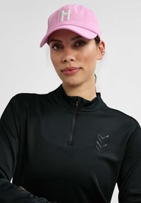 Gorra de béisbol rosa con bordado blanco de la "H"; chaqueta negra con cremallera y un sutil patrón texturizado y detalle de chevron reflectante en el pecho.