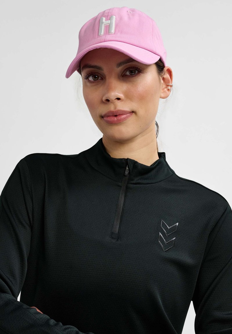 Gorra de béisbol rosa con bordado blanco de la "H"; chaqueta negra con cremallera y un sutil patrón texturizado y detalle de chevron reflectante en el pecho.