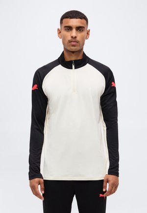 Mand iført en sort og hvid langærmet zip-up top og sorte bukser med røde Puma-logoer på ærmerne og bukserne.
