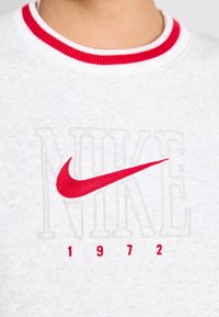 Svijetlosiva dukserica s kragnom u crvenoj i bijeloj prugastoj boji. Sadrži "NIKE" u sivoj boji i crveni Nike swoosh logo, s "1972" ispod u crvenoj boji.