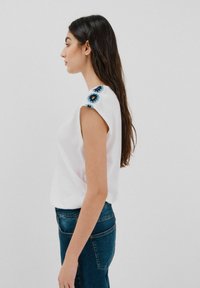 Hvid kortærmet bluse med blomsterbroderi i blå og sort på skulderen, parret med blå denimjeans. Glat stofstruktur.