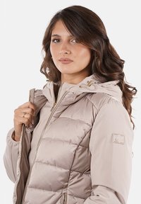 Giacca imbottita color beige chiaro con zip frontale, cappuccio con coulisse e logo ricamato sulla manica. Tessuto liscio e resistente all'acqua.