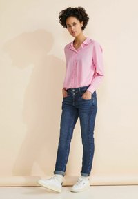 Camisa de manga larga rosa con botones en la parte delantera, combinada con jeans ajustados de color azul oscuro y zapatillas blancas, sobre un fondo neutro.