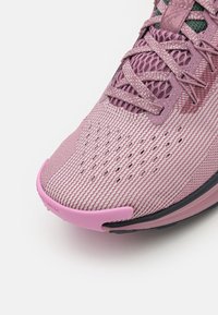 Chaussure de sport rose clair avec une tige en maille texturée. Elle dispose d'une semelle flexible, d'un renfort à l'avant et de détails décoratifs près des lacets.