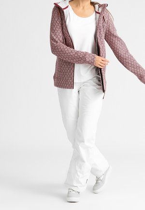 Femme portant une veste zippée rouge à motifs sur un haut blanc, un pantalon blanc et des baskets blanches, posant avec une jambe croisée devant.